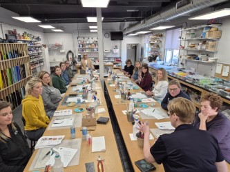 Group of people seated around long tables inside a glass art studio, working with tools and materials, with shelves of glass sheets and supplies visible throughout the room.