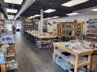 Interior of a glass art studio with worktables, stools, and shelves holding glass sheets, tools, and supplies; large windows and overhead lighting illuminate the workspace.
